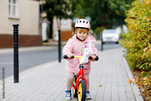 Cute little toddler girl with helmet riding on run balance bike to daycare, playschool or kindergarden. Happy child having fun with learning on learner bicycle. Active kid on cold autumn day outdoors. © Irina Schmidt