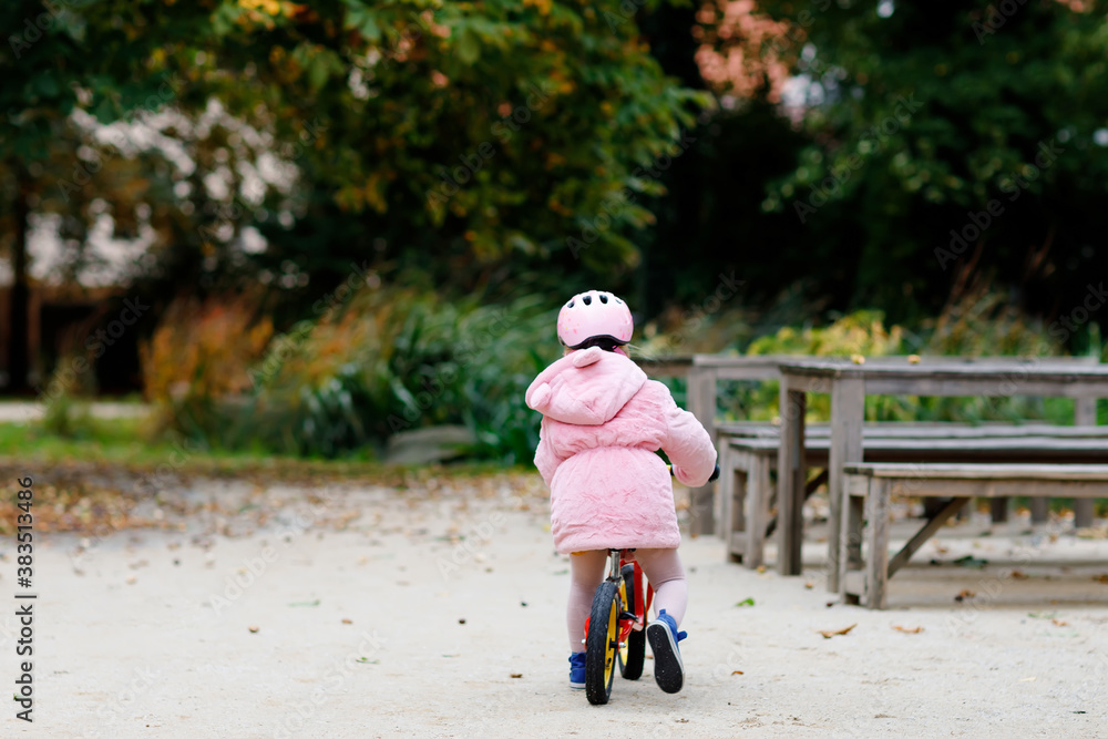 Cute little toddler girl with helmet riding on run balance bike to daycare, playschool or kindergarden. Happy child having fun with learning on learner bicycle. Active kid on cold autumn day outdoors. Cute little toddler girl with helmet riding on run balance bike to daycare, playschool or kindergarden. Happy child having fun with learning on learner bicycle. Active kid on cold autumn day outdoors.