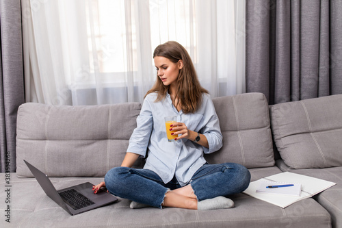 Portrait of a young woman drinking orange juice and surfing in the internet at home sitting on sofa