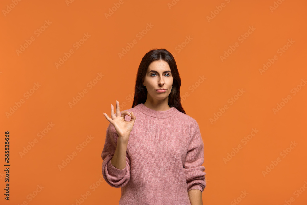 Good job but im not impressed. Not bad. Beautiful woman judging nice work show okay gesture with a neutral expression. wears in pink knitted sweater isolated on orange wall Good job but im not impressed. Not bad. Beautiful woman judging nice work show okay gesture with a neutral expression. wears in pink knitted sweater isolated on orange wall