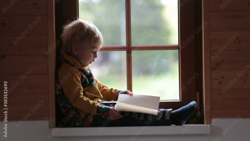 Cute toddler child, blond boy, sitting on window at home, reading book