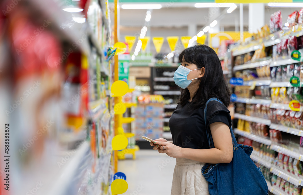Asian women are shopping at the grocery store, holding bphone and wearing a health mask to prevent infection. She's picking goods from the shelves in lifestyle shopping concept,Soft Focus.