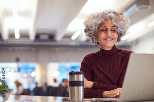 Mature Businesswoman With Travel Mug Working On Laptop In Kitchen Area Of Modern Office