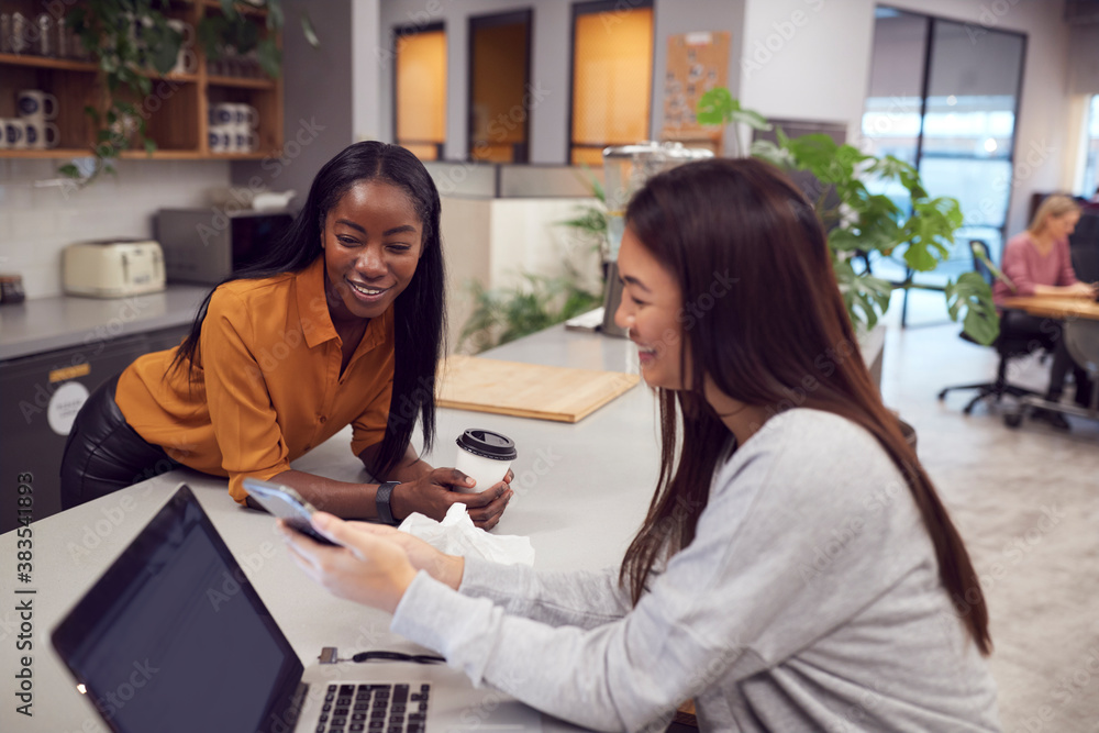 Two Young Businesswomen Working On Laptop In Kitchen Area Of Modern Office Two Young Businesswomen Working On Laptop In Kitchen Area Of Modern Office