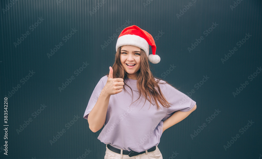 Young stylish trendy woman isolated over grey blue background. Cheerful positive girl in red festive hat hold big thumb up. Like sign or symbol. Winter time for christmas or new year period. Young stylish trendy woman isolated over grey blue background. Cheerful positive girl in red festive hat hold big thumb up. Like sign or symbol. Winter time for christmas or new year period.