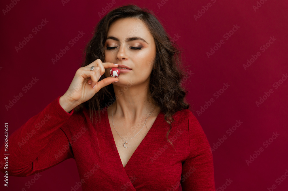 Fototapeta premium Close up of beautiful female holding one round chocolate candy. Woman in red dress posing with little box of bonbons and smelling. Conept of sweet vegan food.
