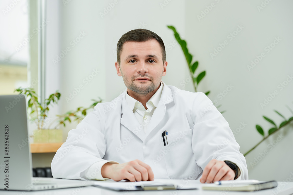 A kind caucasian doctor in a white lab coat is sitting relaxed at his desk and listening in a hospital. A therapist near a laptop is waiting for a patient in a doctor's office.