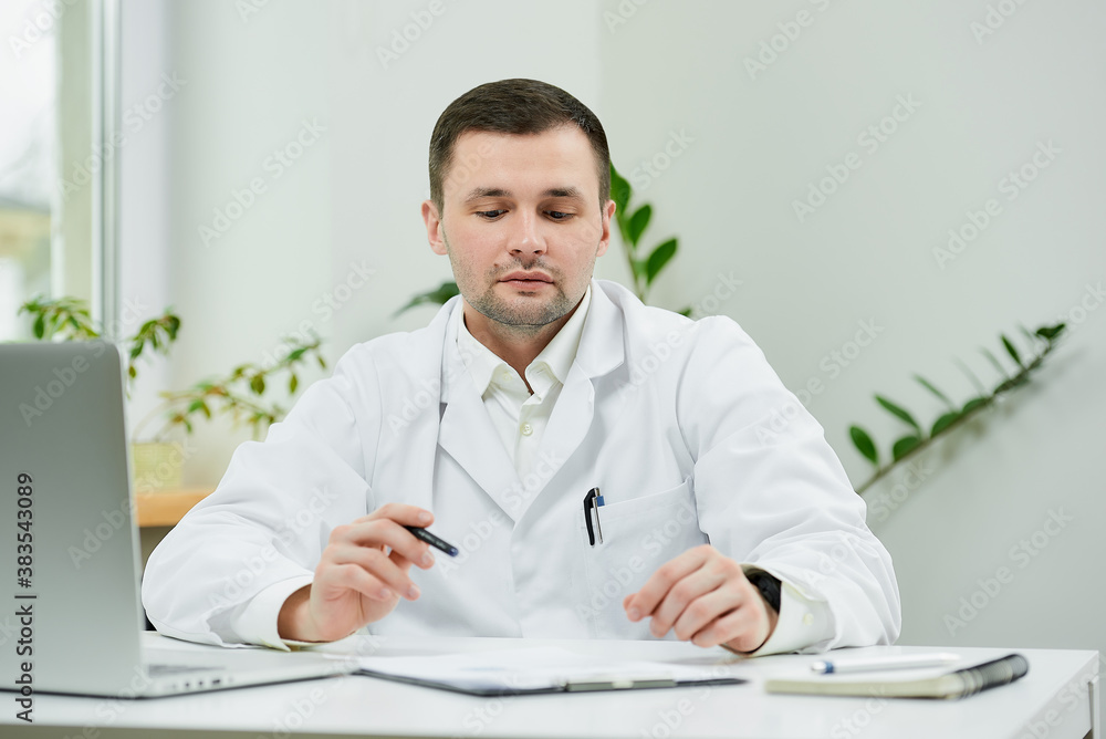 A caucasian doctor in a white lab coat is sitting at his desk and lowered his eyes during examining the patient’s medical history in a hospital. A therapist near a laptop in a doctor's office. A caucasian doctor in a white lab coat is sitting at his desk and lowered his eyes during examining the patient’s medical history in a hospital. A therapist near a laptop in a doctor's office.