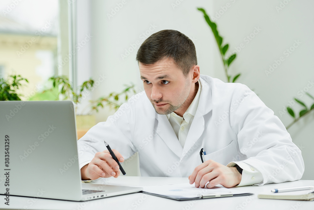 A serious caucasian doctor in a white lab coat is carefully entering the patient’s medical history into a computer in a hospital. A therapist is sitting at a desk with a laptop in a doctor's office.