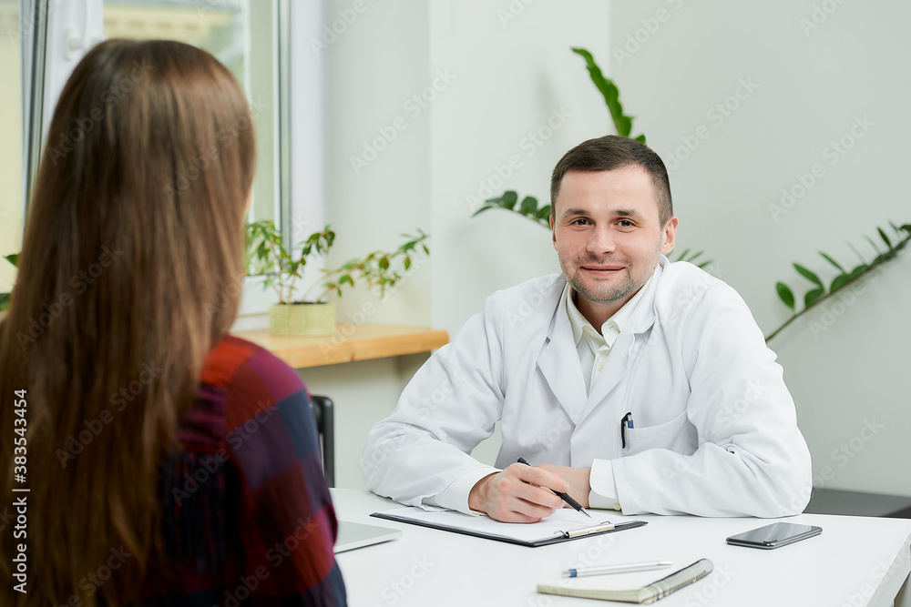 Fototapeta premium A caucasian doctor in a white lab coat is sitting at a desk and listening to a female patient's complaints in a doctor's office. A woman with long hair at a doctor's appointment in a hospital.
