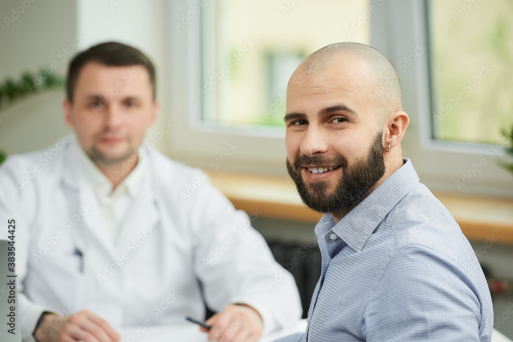 A bald male patient is smiling near a caucasian doctor in a white lab coat in a hospital. A happy man with a beard at an appointment in a doctor's office. A bald male patient is smiling near a caucasian doctor in a white lab coat in a hospital. A happy man with a beard at an appointment in a doctor's office.