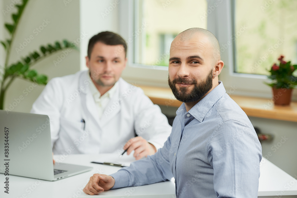 A bald male patient is rejoicing in the success of his healing near a caucasian doctor in a white lab coat in a hospital. A happy man with a beard at an appointment in a doctor's office. A bald male patient is rejoicing in the success of his healing near a caucasian doctor in a white lab coat in a hospital. A happy man with a beard at an appointment in a doctor's office.