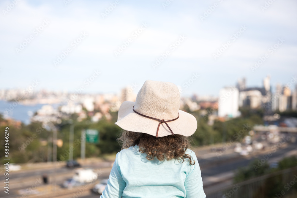 View of a young girl child from behind wearing a broad rim sun hat blue top with her arms outstretched in excitement looking out over the city landscape cityscape Sydney NSW