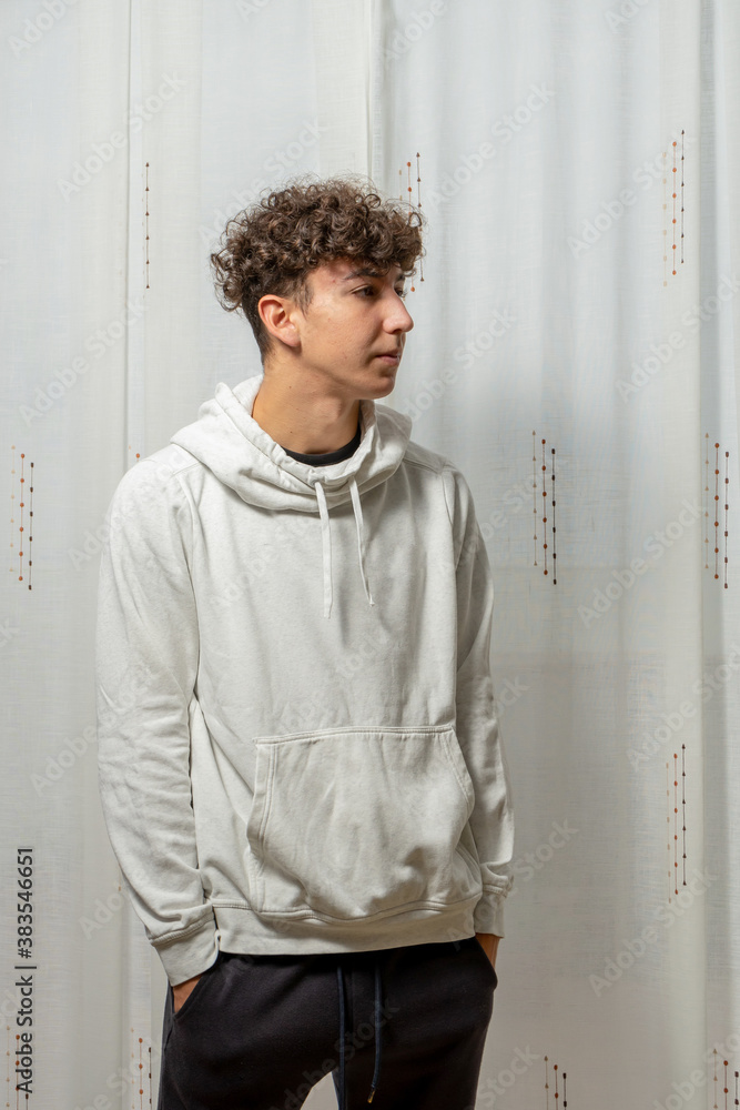Attractive young man with curly hair wearing white sweatshirt posing on white curtains background Attractive young man with curly hair wearing white sweatshirt posing on white curtains background