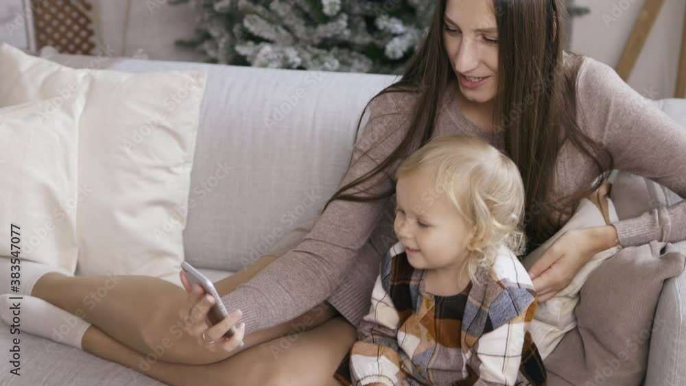 Mother and daughter make phone calls, communicate via video, use a smartphone on Christmas morning. Young mom with her little daughter accept greetings from relatives by the Christmas tree. Mother and daughter make phone calls, communicate via video, use a smartphone on Christmas morning. Young mom with her little daughter accept greetings from relatives by the Christmas tree.