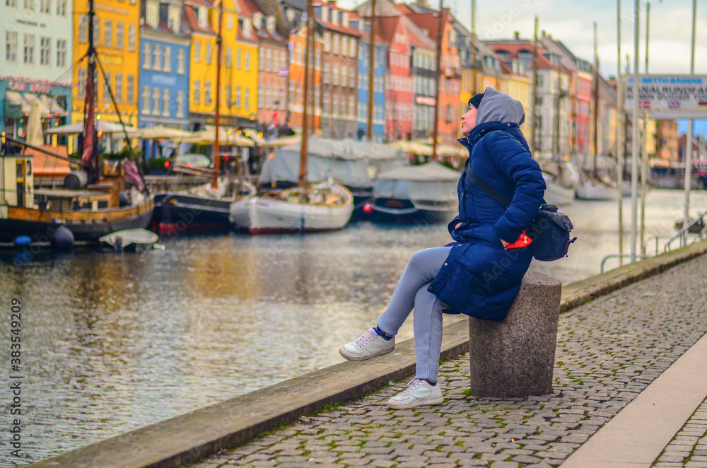 a young woman in warm winter clothes sits and looks at the Nyhavn embankment. Copenhagen, Denmark