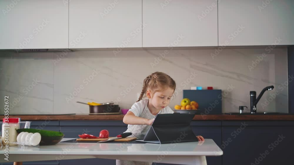 cute little girl in white t-shirt plays on modern tablet at table with cooking ingredients and utensils in large comfortable kitchen at home cute little girl in white t-shirt plays on modern tablet at table with cooking ingredients and utensils in large comfortable kitchen at home