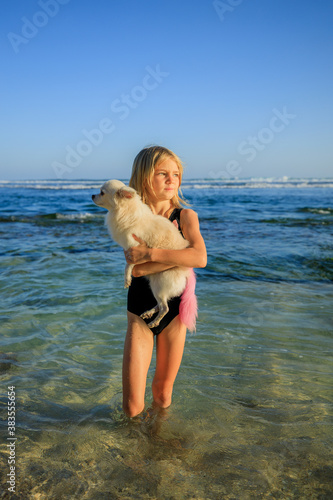 Cute little girl holding small white chihuahua dog with pink tail on the beach. Childhood concept. Spending time outdoor. Ocean during low tide. Bali, Indonesia