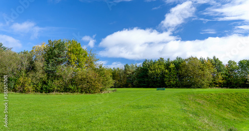 Beautiful natural landscape meadow at public park with could and blue sky in spring or summer. Fresh air panorama view of grass field with metal bench on green forest trees enviroment