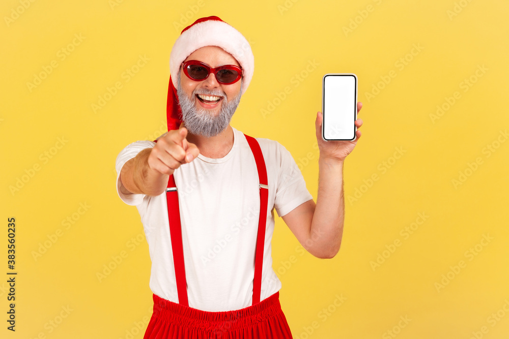 Extremely happy cheerful grey bearded man in santa claus costume pointing finger at camera showing smartphone with empty display, holidays bonuses. Indoor studio shot isolated on yellow background Extremely happy cheerful grey bearded man in santa claus costume pointing finger at camera showing smartphone with empty display, holidays bonuses. Indoor studio shot isolated on yellow background