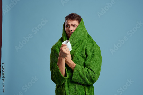 Frozen man in a warm robe with a cup of hot drink on a blue background cropped view