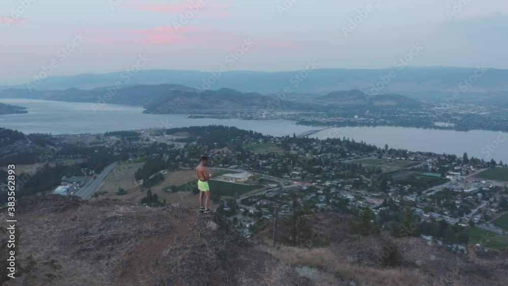 Man stands at top of peak to take photo of mountains, city and lake below Man stands at top of peak to take photo of mountains, city and lake below