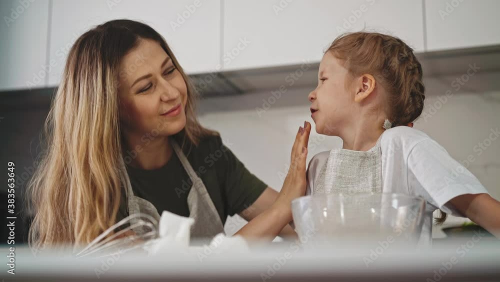 careful blonde mother cleans smiling little daughter chin by hand cooking together at table in light modern kitchen at home careful blonde mother cleans smiling little daughter chin by hand cooking together at table in light modern kitchen at home