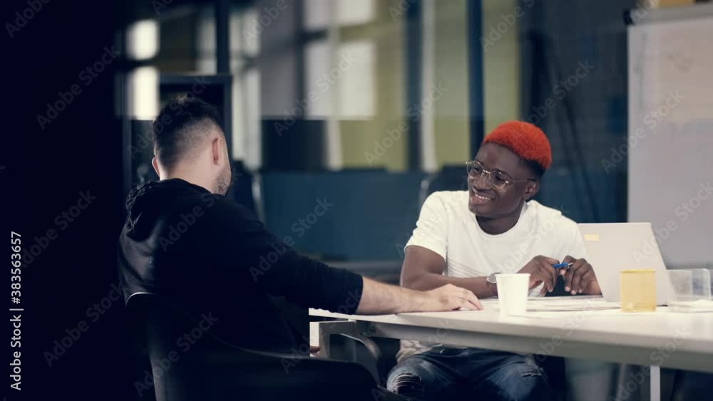 Two male colleagues have a positive business conversation discussing creative ideas. An African American businessman listens attentively to a colleague and rejoices at promising opportunities for the Two male colleagues have a positive business conversation discussing creative ideas. An African American businessman listens attentively to a colleague and rejoices at promising opportunities for the