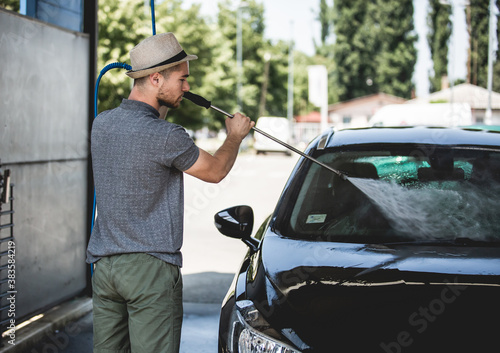 Young man cleaning automobile with high pressure water jet at car wash.