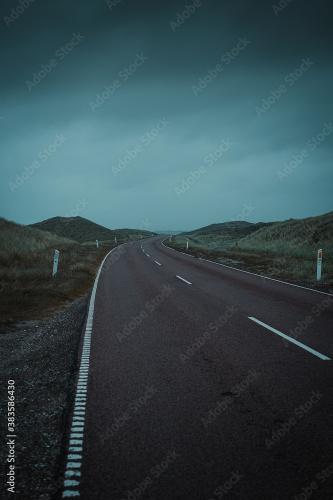 A curved road trough the wide beach dune landscape on a dramatic moody dark and rainy storm day at the ocean. Endless driving roads in the National Park Thy in Denmark