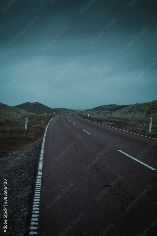 Naklejka premium A curved road trough the wide beach dune landscape on a dramatic moody dark and rainy storm day at the ocean. Endless driving roads in the National Park Thy in Denmark