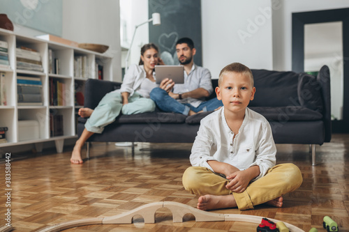young boy sitting on floor and playing with wooden train toy, mother and father sitting on sofa in blurred background
