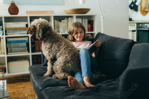 girl sitting on sofa reading book with her dog at home