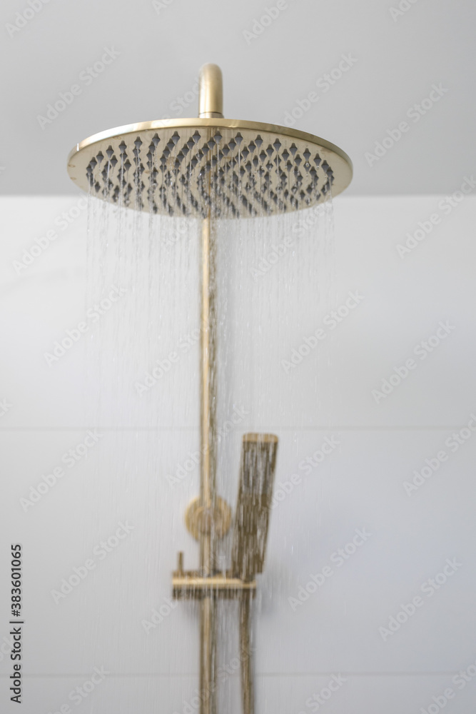 Brushed brass rain mixer shower head with running water isolated against white tiled wall in a new modern elegant bathroom lit by natural light from a nearby window modern interior house renovation 