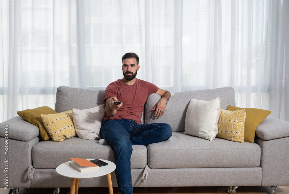 Young casual man sitting on the sofa at his home with remote control in his hand relaxing with television program Young casual man sitting on the sofa at his home with remote control in his hand relaxing with television program