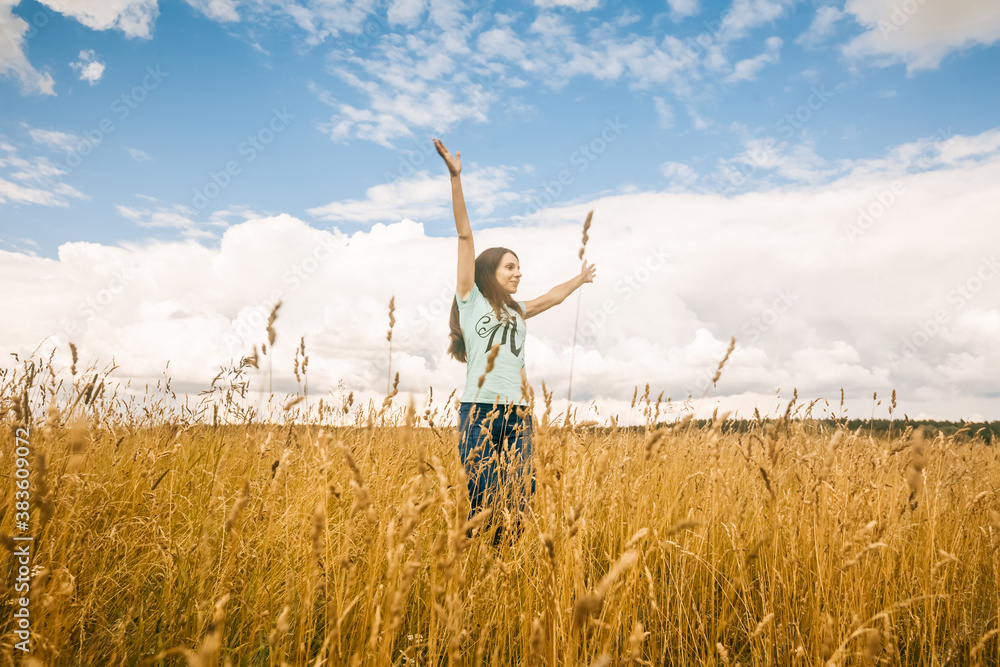 Portrait of young woman walking on field. Girl jump with raised hands. Nature beauty, blue sky,white clouds and field with golden wheat. Outdoor lifestyle. Freedom concept