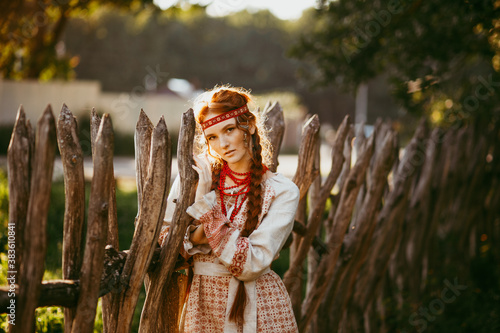 A beautiful Slavic girl with long blonde hair and brown eyes in a white and red embroidered suit stands by a wooden fence.Traditional clothing of the Ukrainian region.