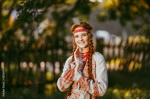 A beautiful Slavic girl with long blonde hair and brown eyes in a white and red embroidered suit stands by a wooden fence.Traditional clothing of the Ukrainian region. © Aleksei Zakharov