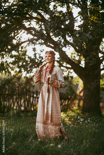 A beautiful Slavic girl with long blonde hair and brown eyes in a white and red embroidered suit stands by a wooden fence.Traditional clothing of the Ukrainian region.
