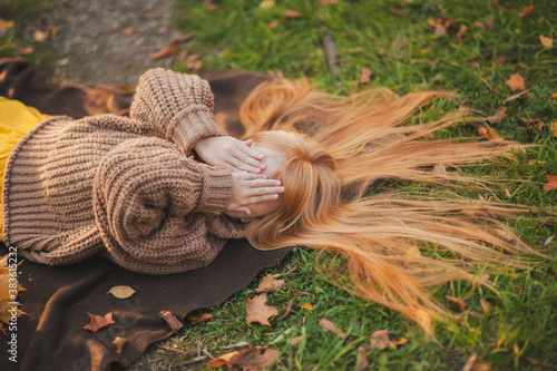 A little girl in a yellow dress and a brown sweater lies on a brown blanket in an autumn Park. Autumn photo shoot of a girl with red hair.