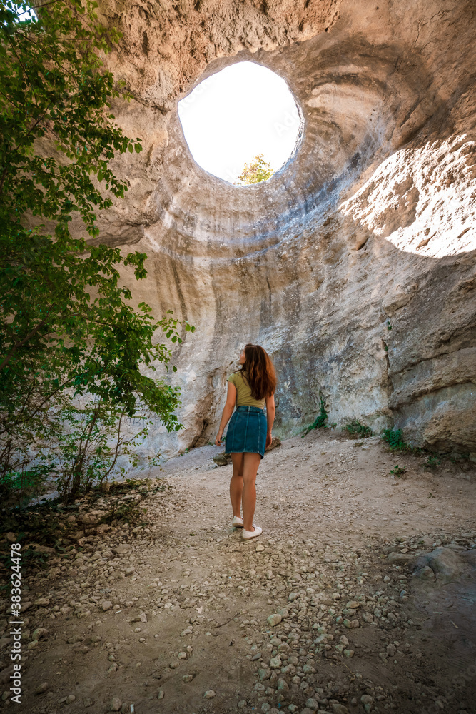 Rear view of a young woman adventurer standing in an underground cave with an opening to the outside, Sunlight through a cave