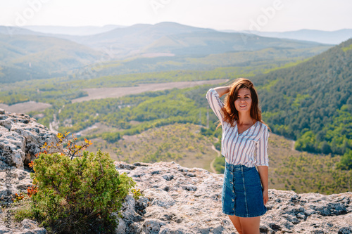 A beautiful woman stands on a high cliff, enjoying the view of the mountain range and forest © KseniaJoyg