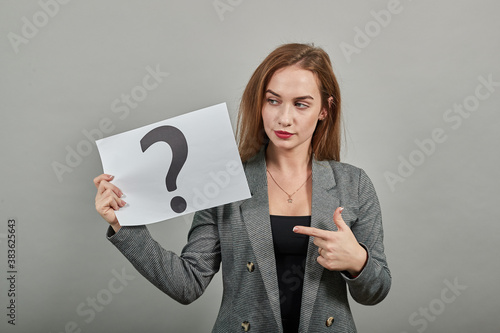 Black question mark on a piece of white paper. Close-up hand holding symbol incomprehensibility. Showing sign ignorance near face isolated in studio. Young attractive woman, dressed gray jacket photo