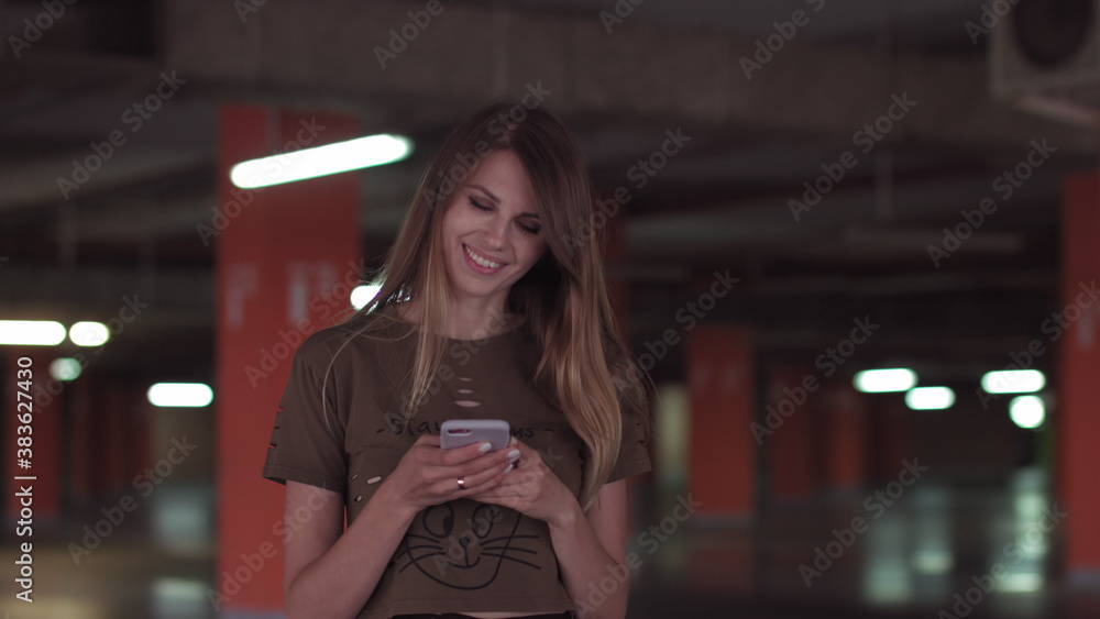 Relaxed young woman using smartphone surfing social media, checking news, playing mobile games or texting messages standing in the supermarket parking lot.