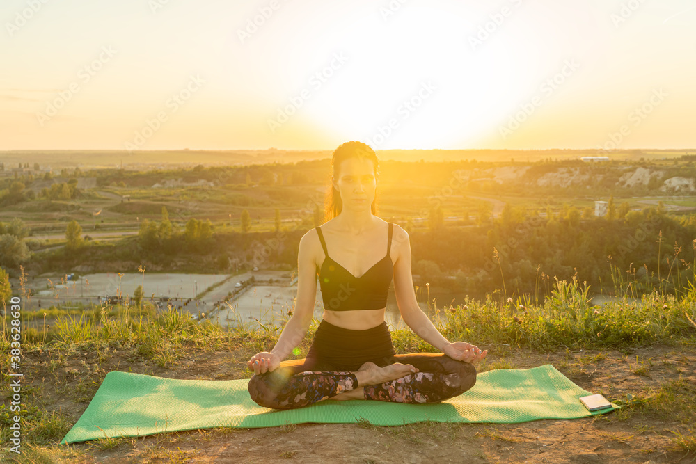 Young woman meditating in lotus pose on nature at sunset.