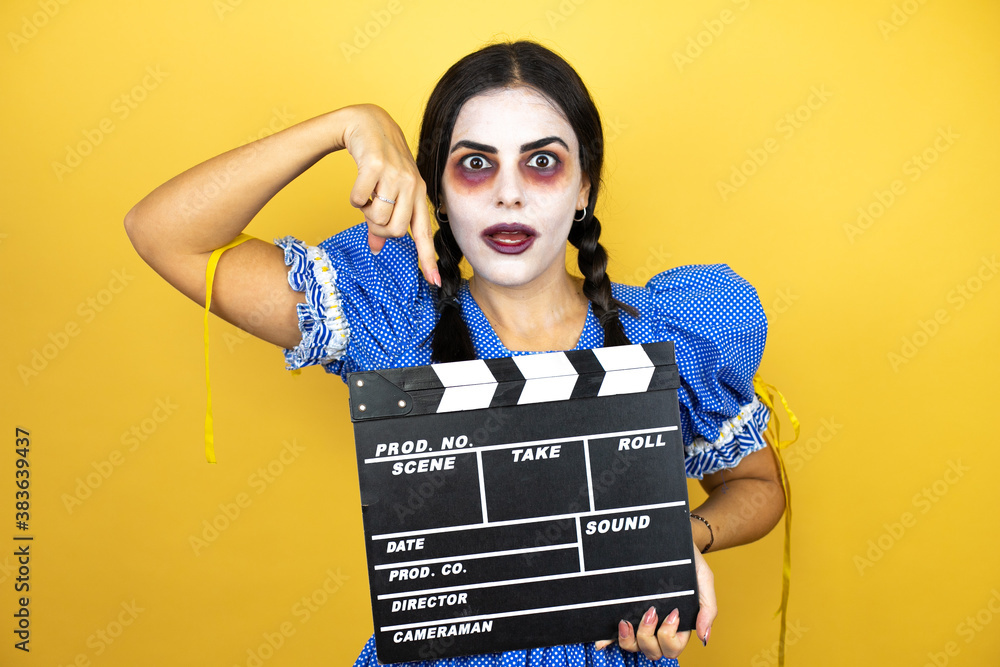 woman wearing a scary doll halloween costume over yellow background holding clapperboard very happy having fun