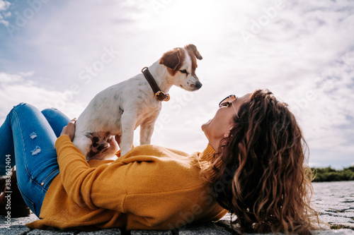A beautiful little dog sitting on its owner looking at her face. The woman is lying down in the park in a sunny day in Madrid. Family dog outdoor lifestyle photo