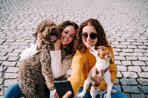 Photography Two beautiful women sitting in the street with their pets and taking a photo with a phone