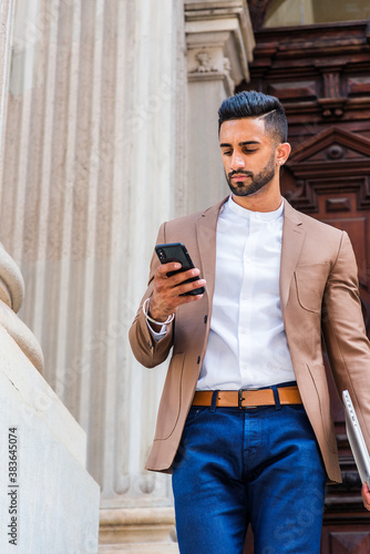 Obraz na płótnie Young East Indian American Man with beard, wearing brown blazer, white round collar shirt, blue pants, carrying laptop computer, standing by old style doorway, looking down, texting on cell phone.. w salonie