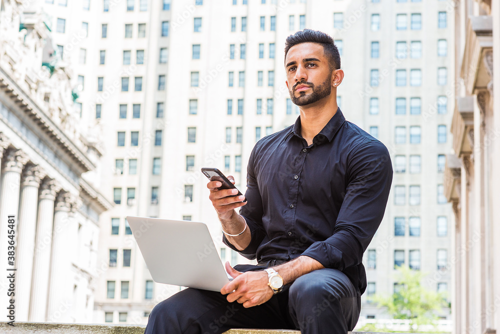Young East Indian American Businessman with beard traveling, working in New York City, wearing black shirt, holding laptop computer, sitting outside old style office building, texting on cell phone..